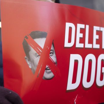 A protestor holds a sign that has a picture of Elon Musk with an X over his face, and text that says "Delete DOGE"