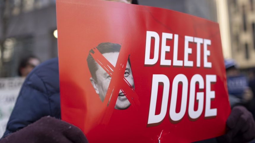 A protestor holds a sign that has a picture of Elon Musk with an X over his face, and text that says "Delete DOGE"
