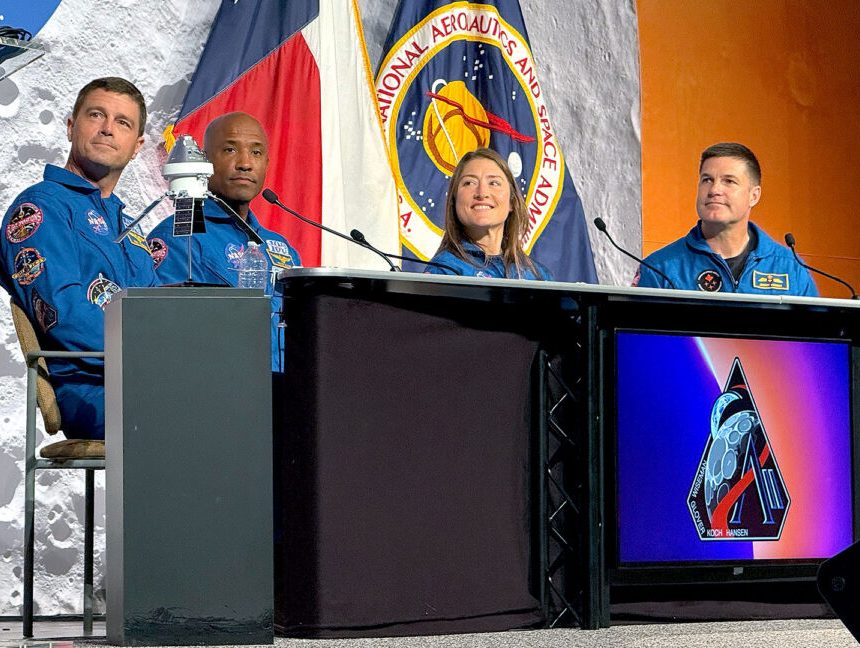 Three men and a woman, all in blue flight suits, sit at a desk during a press conference
