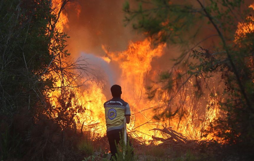 Image of a lone person with a hose, standing before a wall of flame.