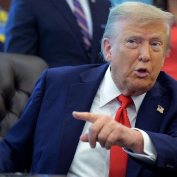President Donald Trump sits at his desk and talks while pointing a finger.