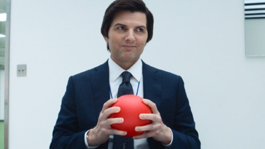 man in a suit in a bland office setting holding a bright orange-red ball in his hands.