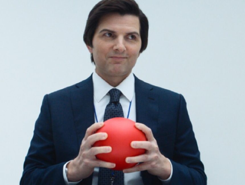man in a suit in a bland office setting holding a bright orange-red ball in his hands.