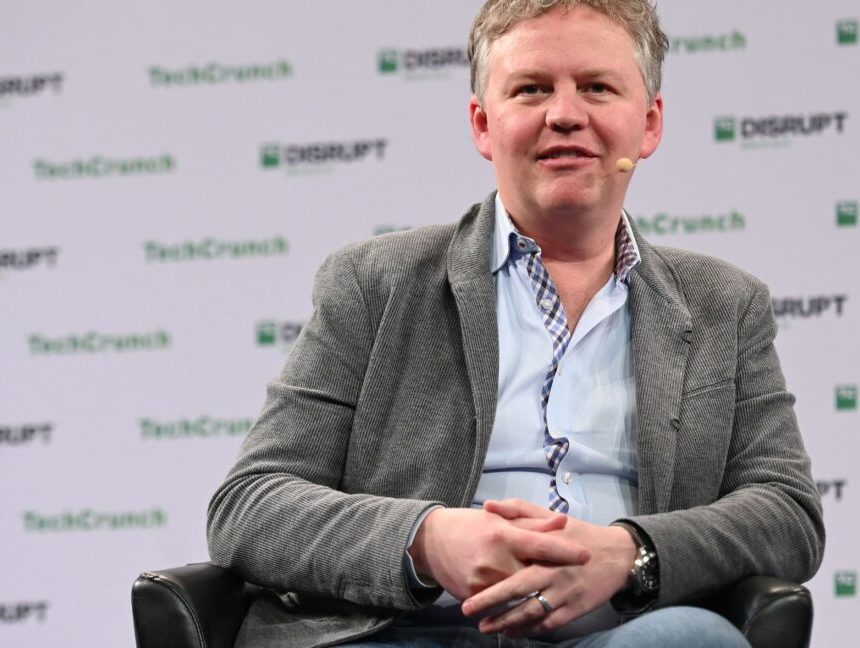 A man in a suit jacket sits at a TechCrunch speaking event