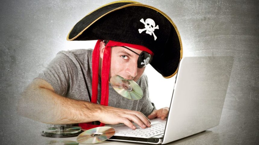 A man, surrounded by music CDs, uses a laptop while wearing a skull-and-crossbones pirate hat and holding one of the CDs in his mouth.