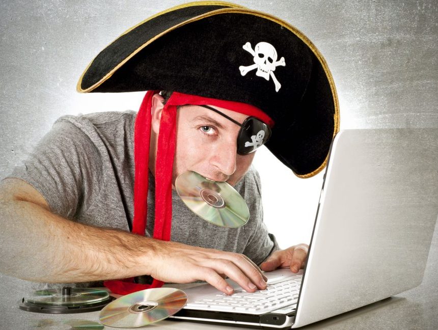A man, surrounded by music CDs, uses a laptop while wearing a skull-and-crossbones pirate hat and holding one of the CDs in his mouth.