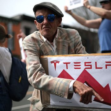 Tesla protester holds a sign showing Tesla's stock price going down.