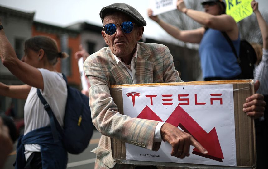 Tesla protester holds a sign showing Tesla's stock price going down.