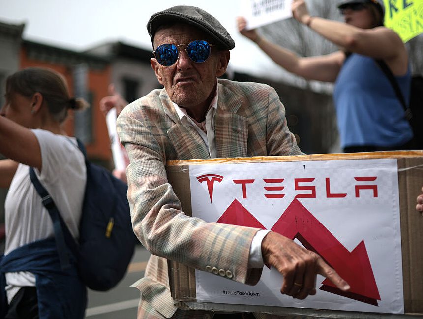 Tesla protester holds a sign showing Tesla's stock price going down.