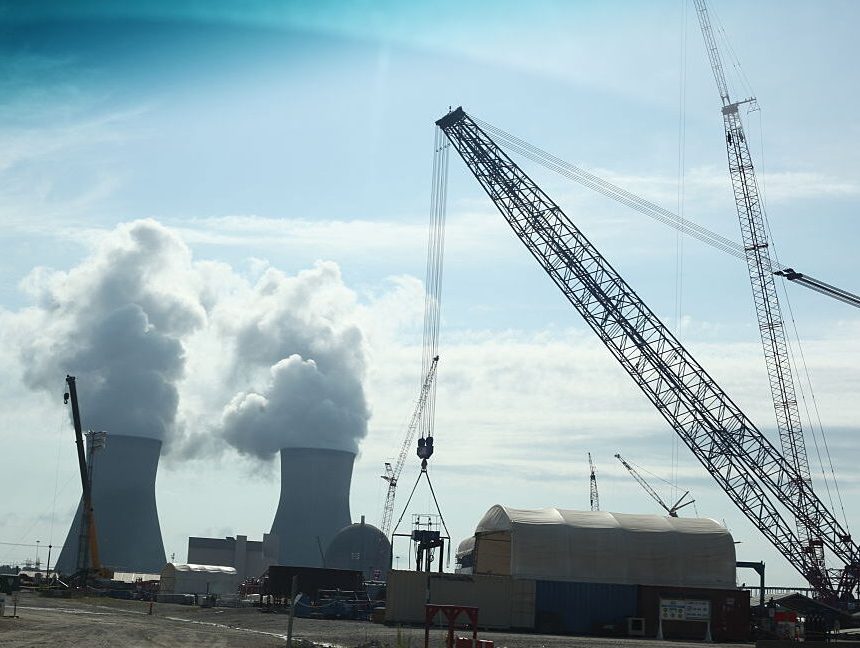Construction cranes in action in the foreground, while steam spills from two cooling towards in the background.