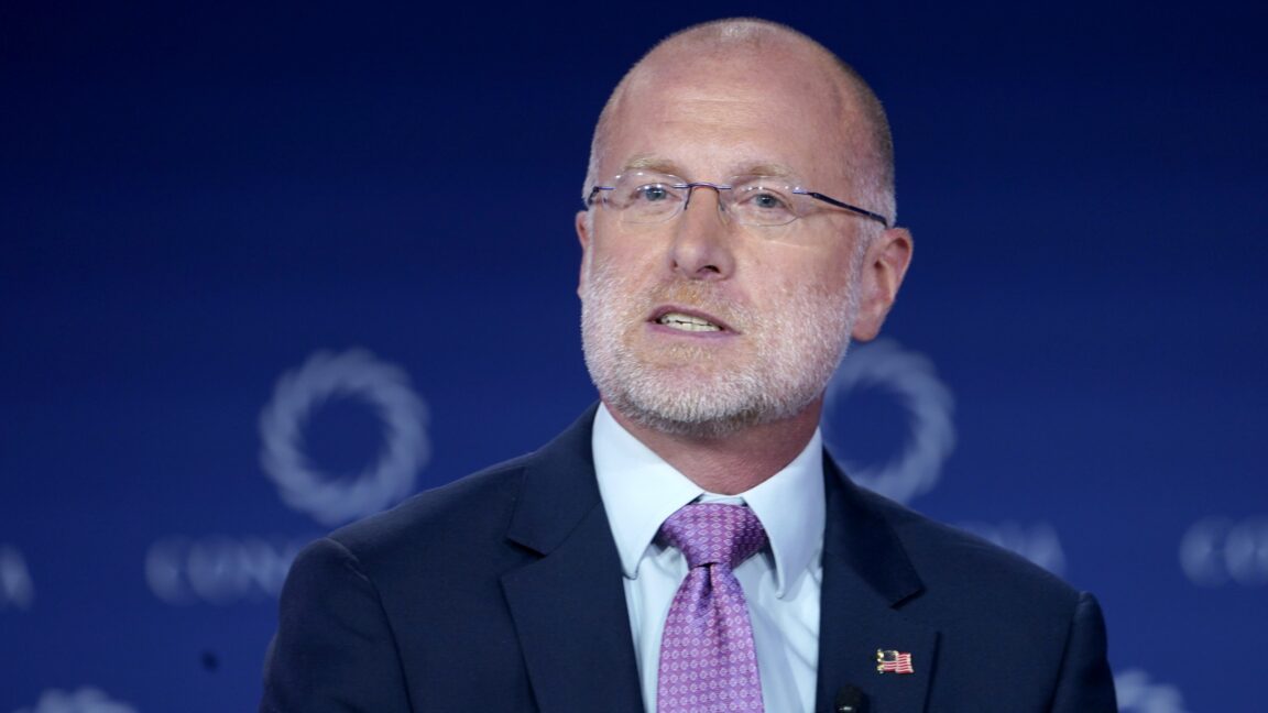 FCC Chairman Brendan Carr speaking on stage at a conference, wearing an American flag pin and a microphone clipped to his suit jacket.