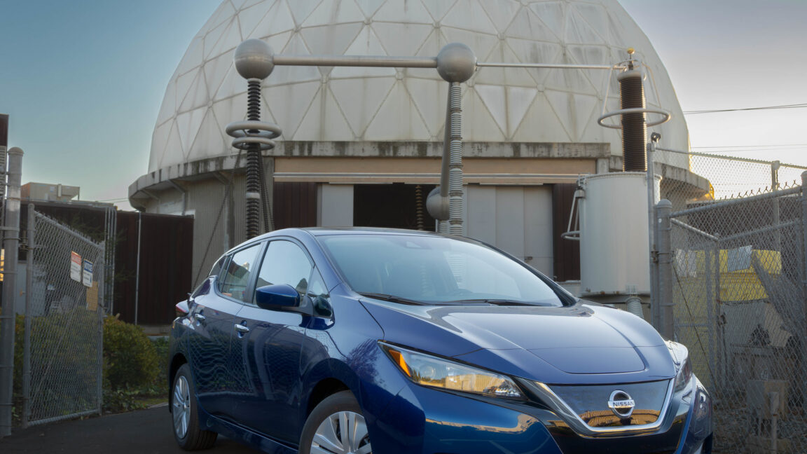 A blue Nissan Leaf in front of some electrical equipment and what looks like a concrete dome