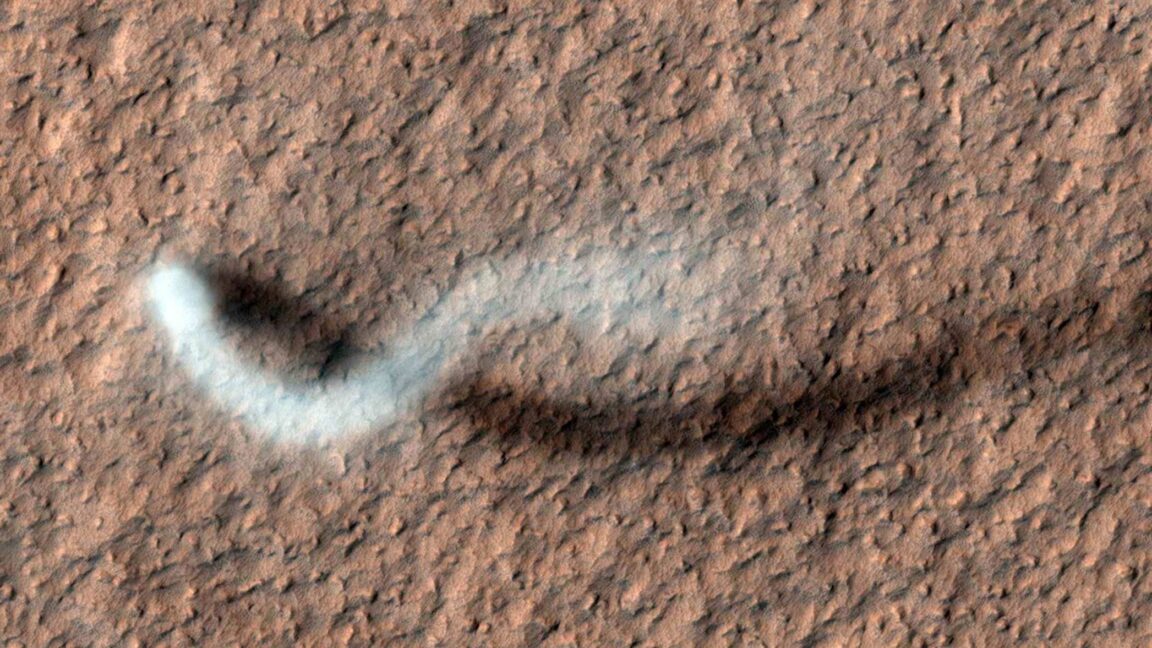 Image of a rough, hilly landscape taken from above. The landscape is barren and reddish. At the left , the white plume of a dust devil stretches upwards before diffusing off to the right.