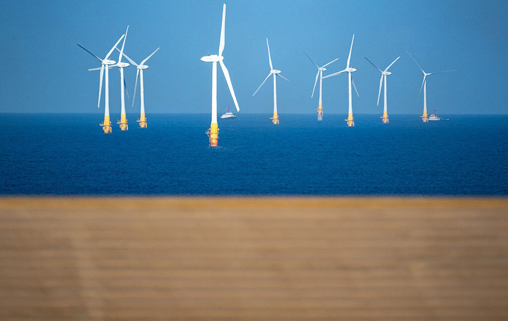 Image of a series of wind turbines standing in blue waters off a golden sand beach.