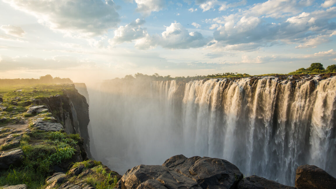 a view of a broad waterfall, with mist rising above rocks in the foreground.